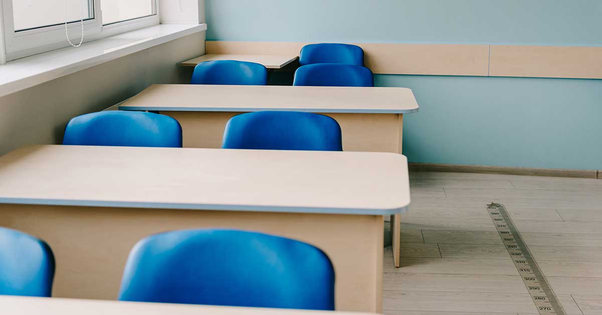 Empty classroom with light wooden desks, blue plastic chairs, and a window on the left. A metal ruler lies on the floor in the foreground, evoking stories of learning and migration shaped by immigration.