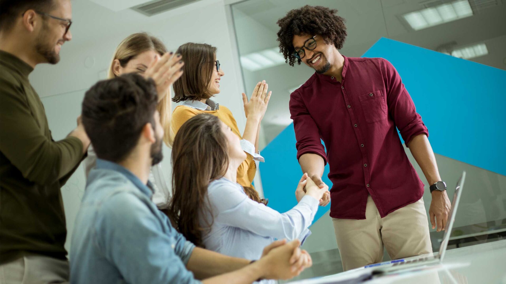 A group of colleagues in an office stands and sits around a table, smiling and clapping as one workforce member shakes hands with another.