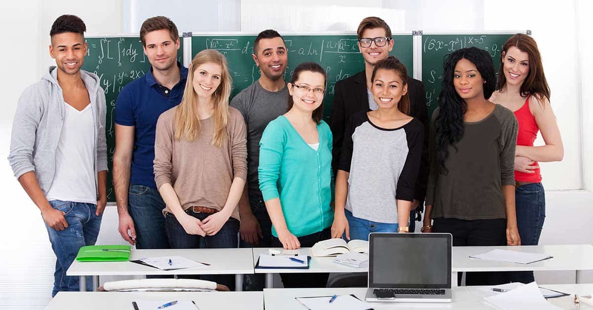 A diverse group of young adults stands in a classroom in front of a chalkboard filled with mathematical equations, embodying the spirit of college prep as they smile for the camera.