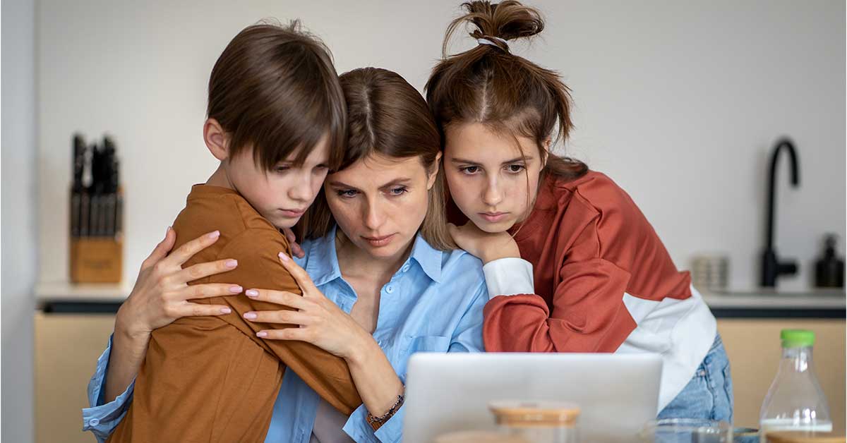A woman sits at a table with two children, all looking seriously at an online video on a laptop screen, with the woman embracing the children closely.