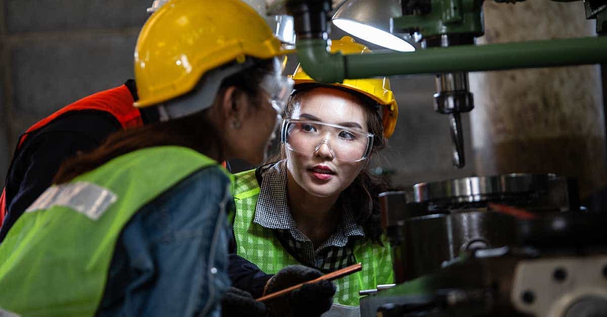 Two workers in safety gear and hard hats discuss near machinery in an industrial setting.