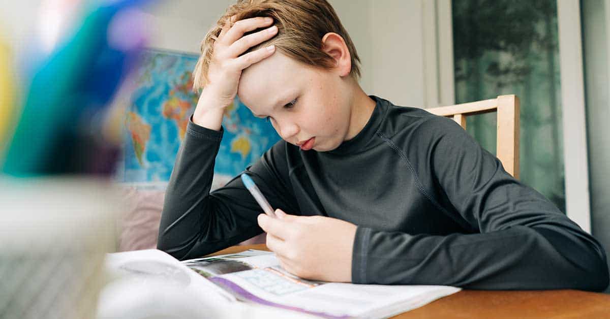 A boy sits at a table, holding a pen and looking intently at an open book, appearing focused while working on his homework.