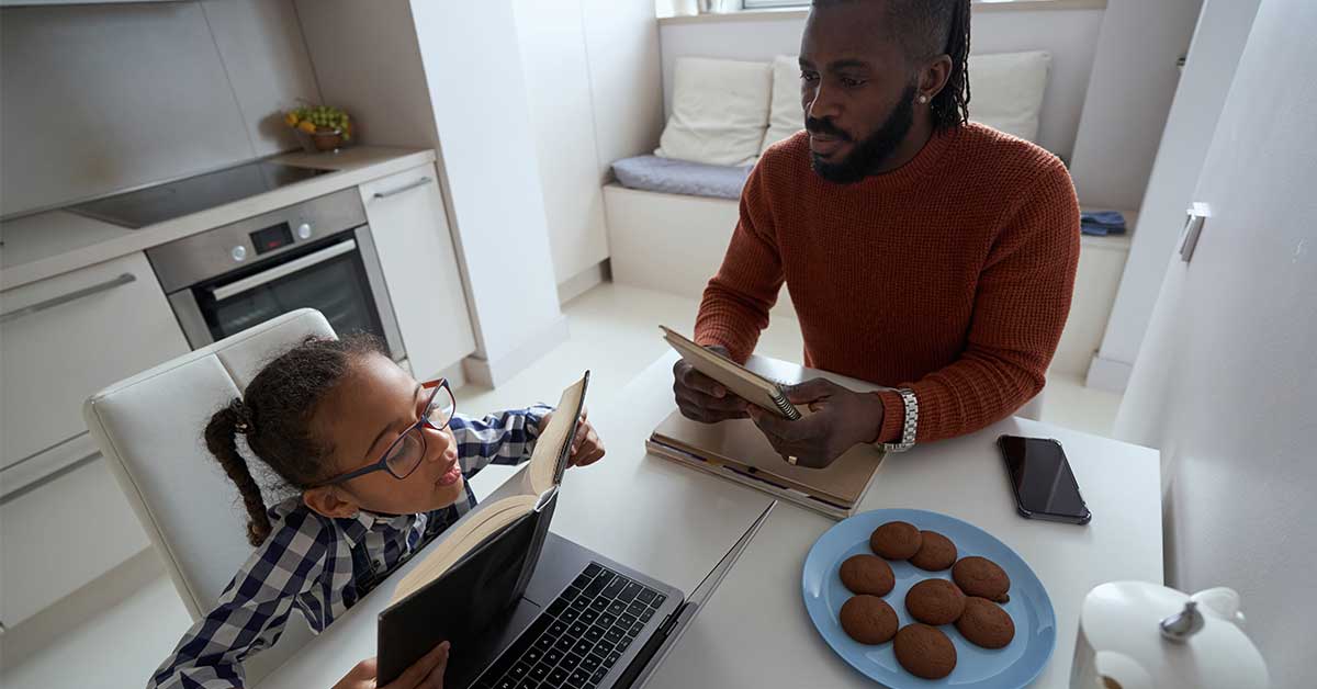 A man and a child sit at a kitchen table with books and a laptop, homeschooling together, while a plate of cookies and a phone are on the table.
