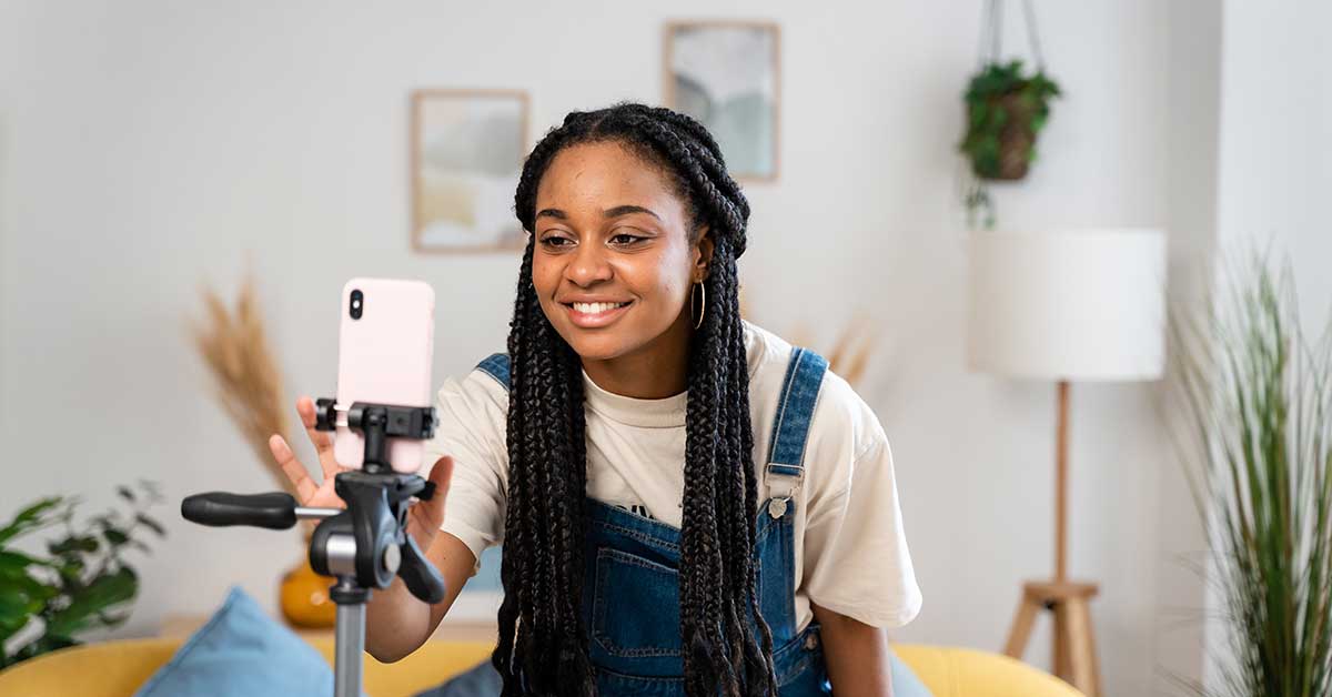 An influencer with long braids smiles at a smartphone mounted on a tripod while sitting in a living room.