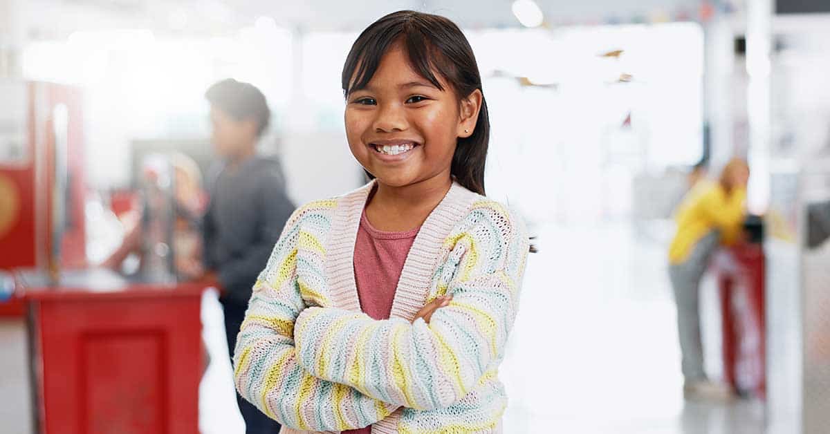 Smiling young student with arms crossed stands indoors, wearing a striped sweater and pink shirt; blurred students and bright background visible.