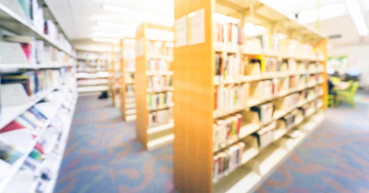 A blurred image of a library interior with wooden bookshelves filled with books and a patterned carpet, echoing the silent collapse of American public education.