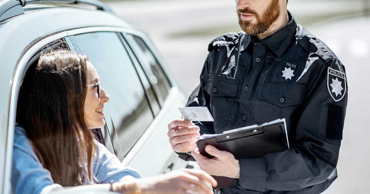 A police officer holds a clipboard and examines a driver's license handed to him by a woman sitting in her car, discussing sovereign identification.