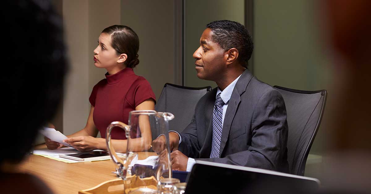 Two people sit at a conference table, engaged in a governance discussion. One is wearing a suit, the other a maroon top. Both listen attentively, with papers and a glass pitcher on the table.