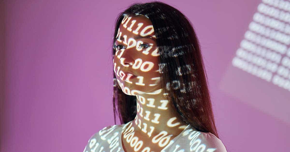 A woman with long dark hair is illuminated by projected binary code, embodying AI predictions against a vibrant pink background.