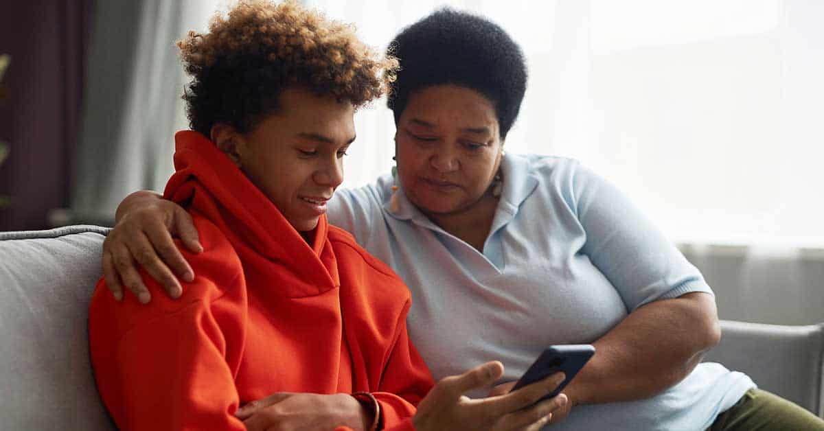 A woman sits on a couch with her arm around a child in a red hoodie as they look at a smartphone together.