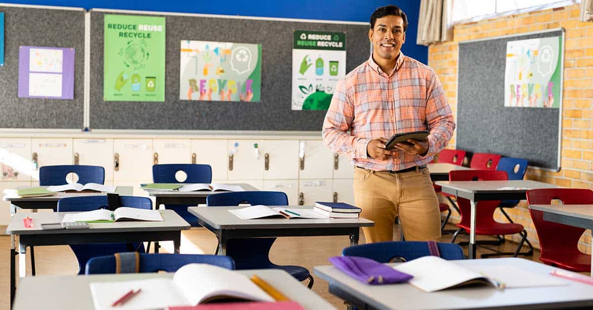 A man stands in a classroom holding books. Desks feature open books and stationery, while posters on the wall promote recycling and highlight tribal sustainable systems.