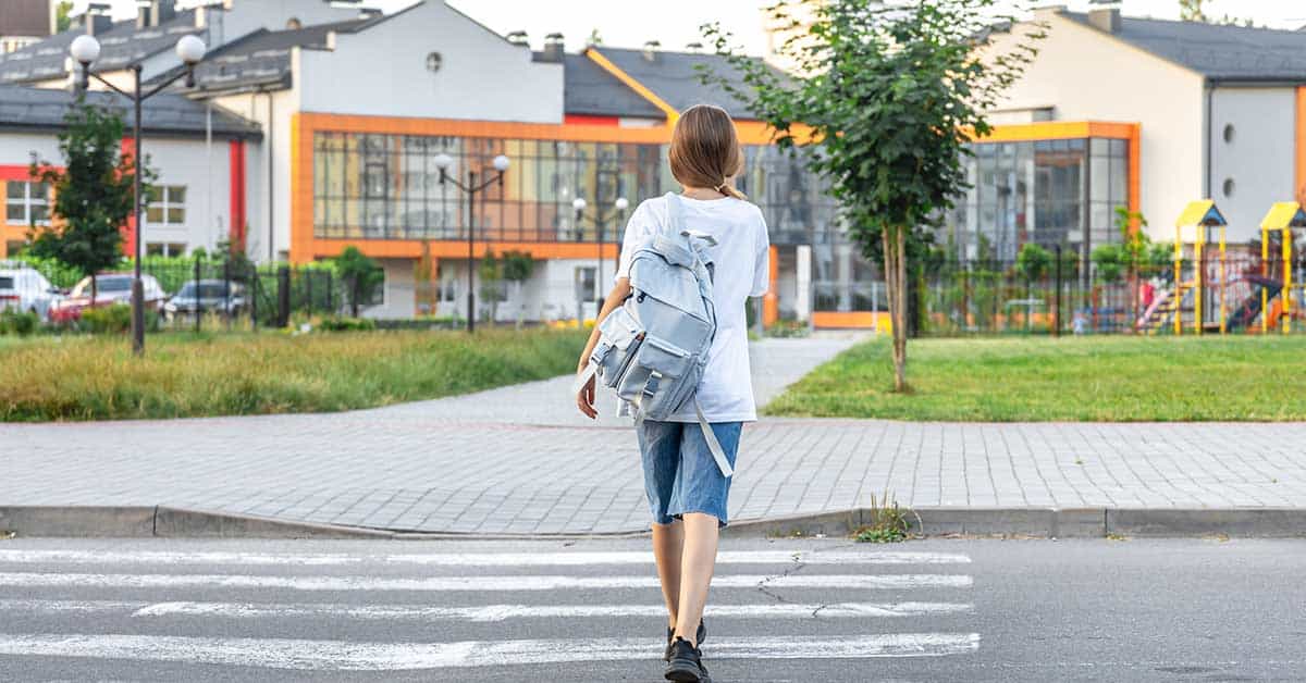 A student with a backpack crosses a street toward a modern school building on a sunny day, unaware of the recent executive order impacting local education.