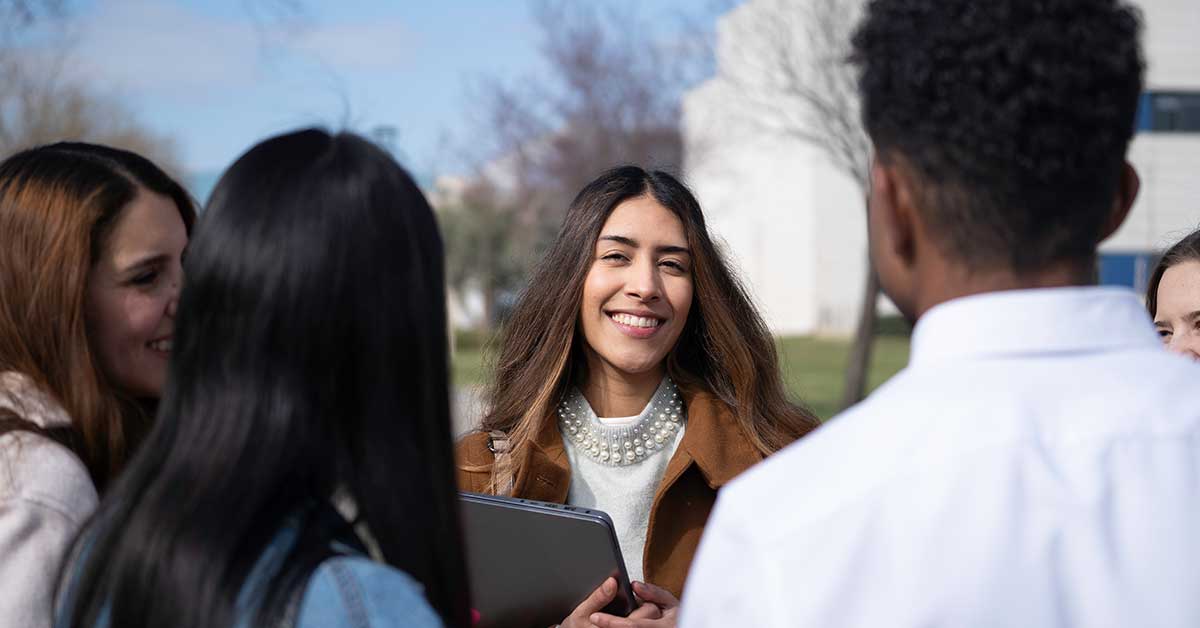A group of young adults stand outdoors talking, with one woman in the center smiling and holding a laptop, discussing their recent BIE project.