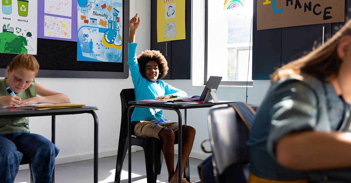 A student sits at a desk in a classroom raising his hand in compliance, while other students work at their desks. Educational posters are visible on the wall.