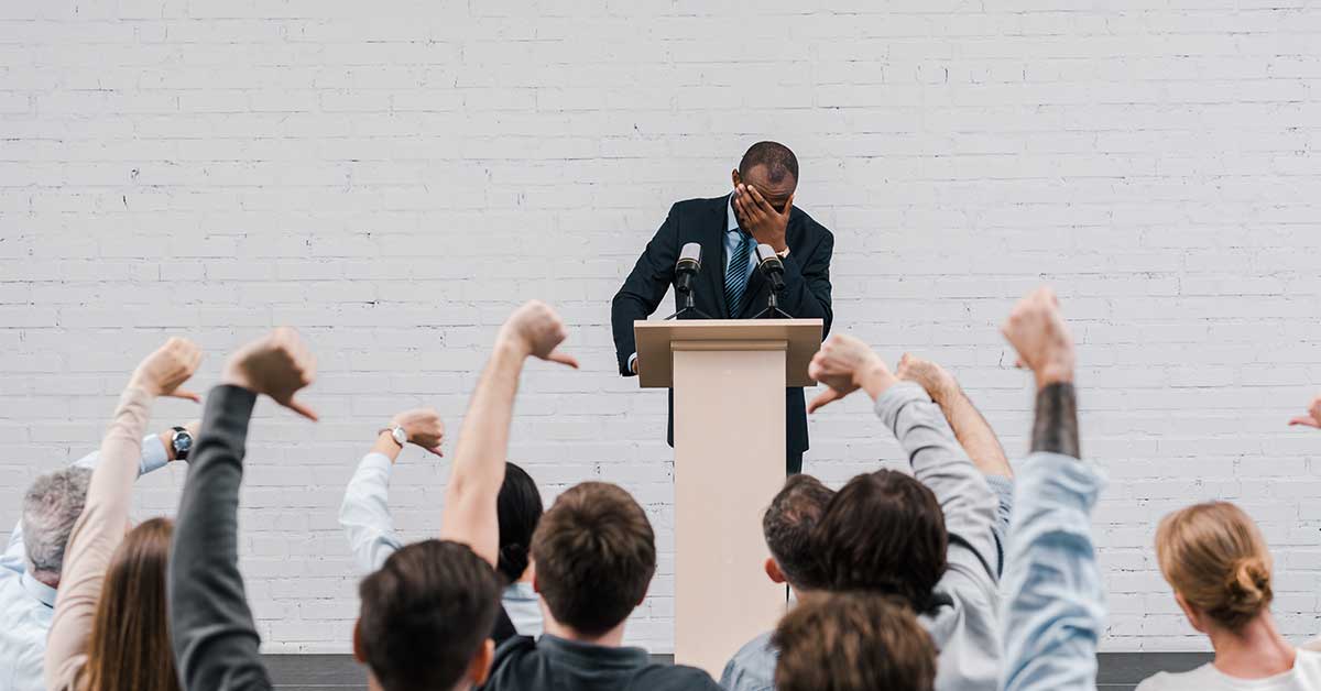 A man in a suit stands at a podium with his head in his hand, facing an audience giving thumbs down gestures—highlighting the challenges and pressures faced in education.