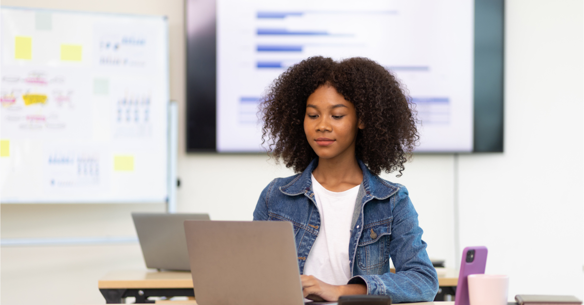 A woman with curly hair works on a laptop in a classroom or office, analyzing AI charts displayed on a screen, with a whiteboard in the background.