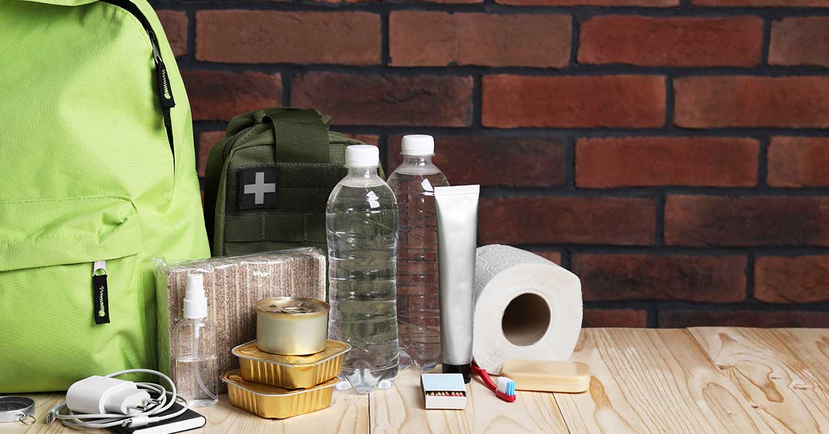 Emergency supplies for a wildfire, including a green backpack, first aid kit, water bottles, canned food, bandages, matches, toothpaste, toothbrush, soap, and toilet paper on a wooden table.