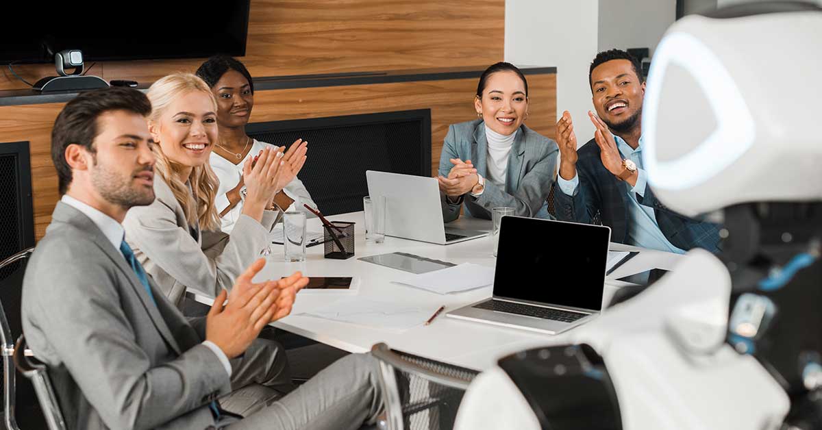 Five business professionals sit at a conference table, clapping and smiling at a humanoid AI robot during a meeting. Laptops and papers are on the table.