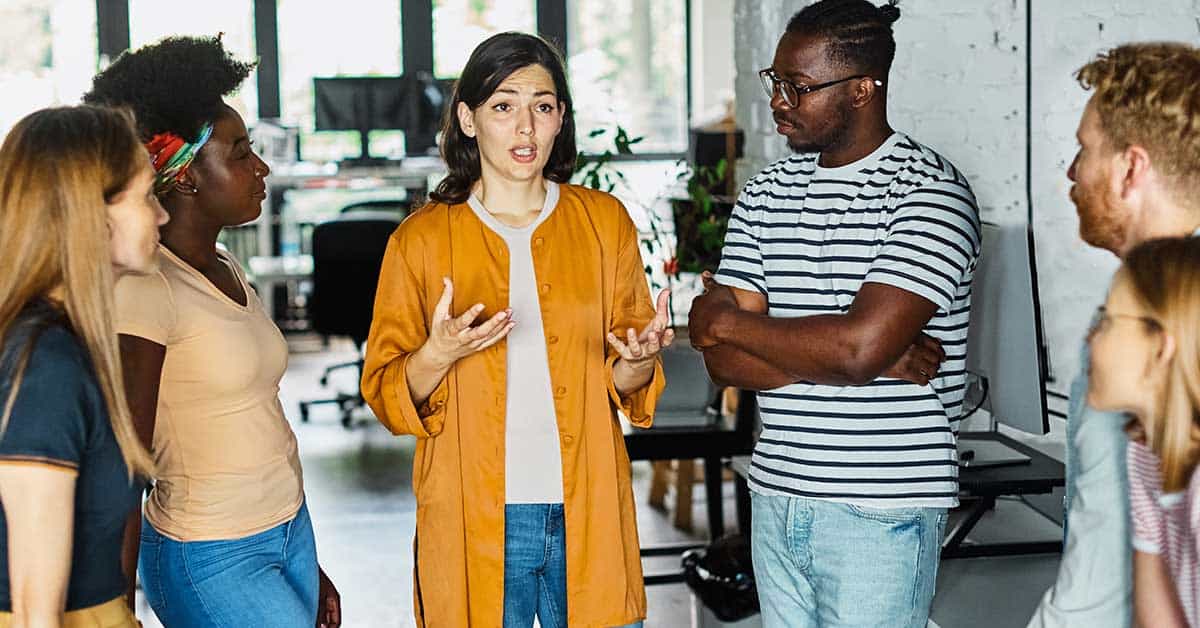 A woman in an orange jacket speaks to a group of five people, as employers gather in a casual office setting.