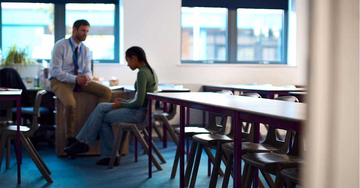 A teacher and student discuss progressive discipline in a bright classroom with large windows, a row of tables, and a plant, fostering a supportive learning environment.