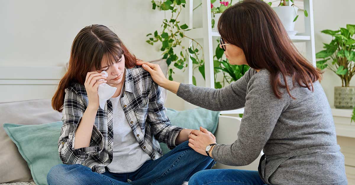 A woman comforts a crying young woman sitting on a couch, offering support by touching her shoulder and knee after she reveals she's been affected by sextortion.