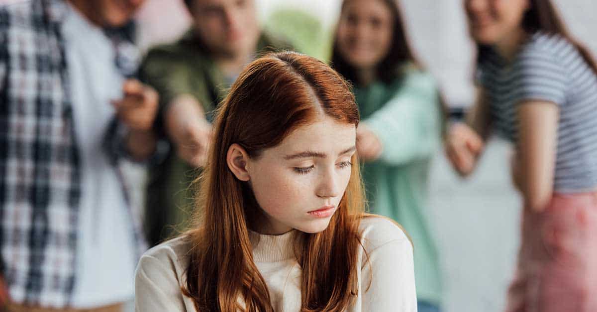 A girl with red hair looks down sadly while a group of peers in the background point at her and laugh, having been tricked by a deepfake.