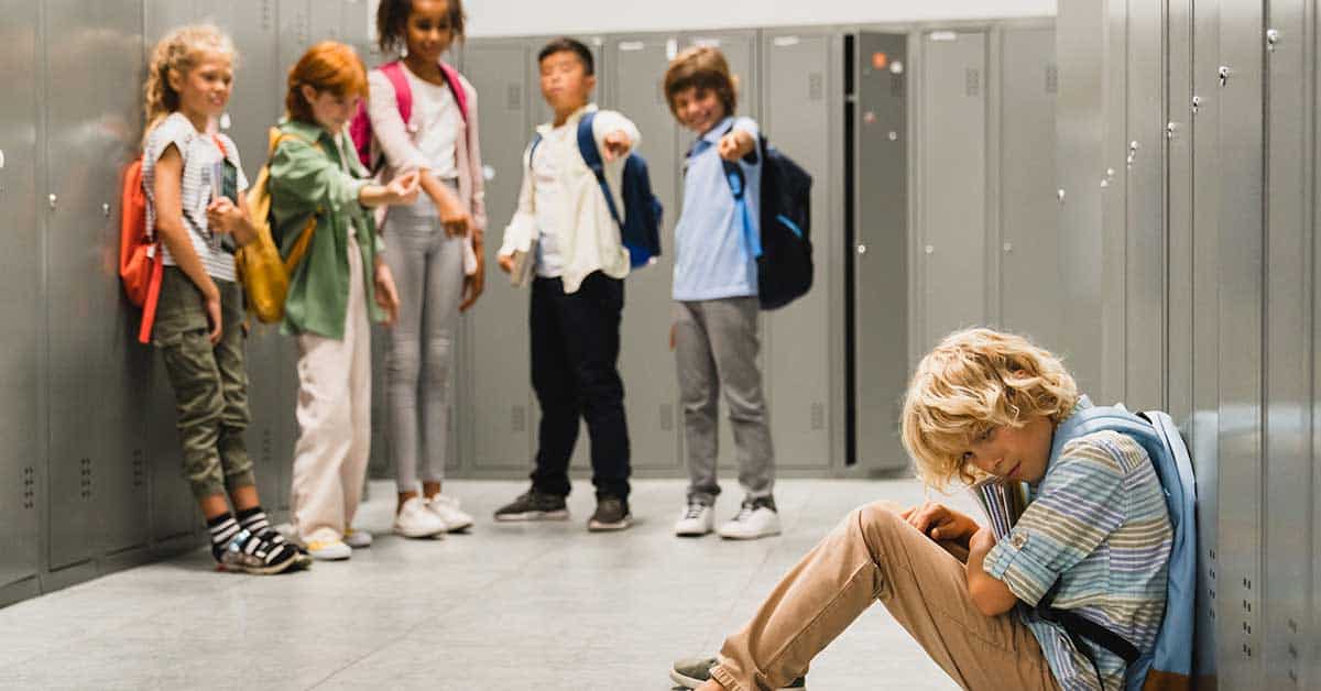 A group of children in a school hallway point and laugh at a blonde boy sitting alone on the floor, looking upset near the school lockers.