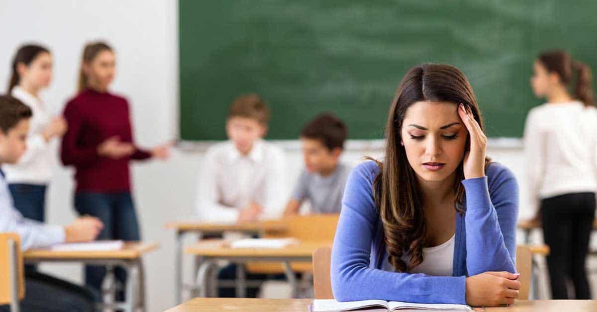 A person sits at a desk appearing stressed, with an open book in front. In the background, a group of people are discussing the teacher shortage crisis. A chalkboard is visible on the wall.