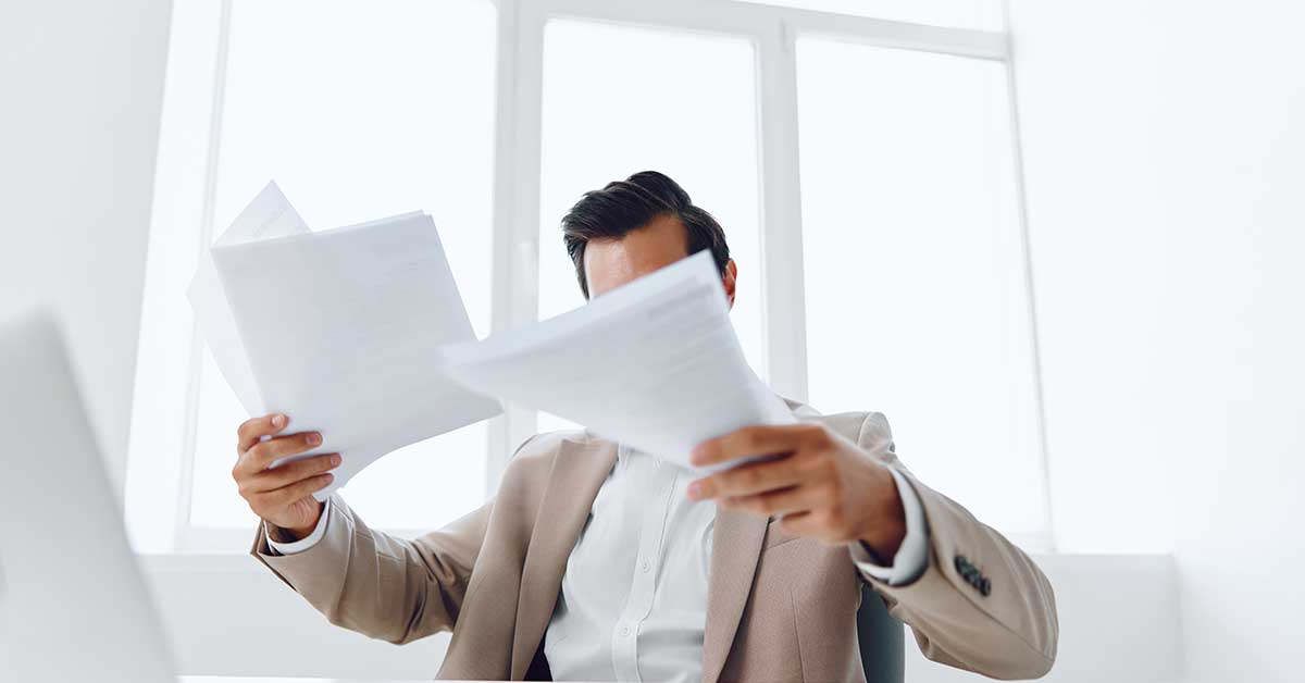 A person in a suit is holding and reviewing multiple sheets of paper marked with "Confidentiality" at a desk in a bright office.