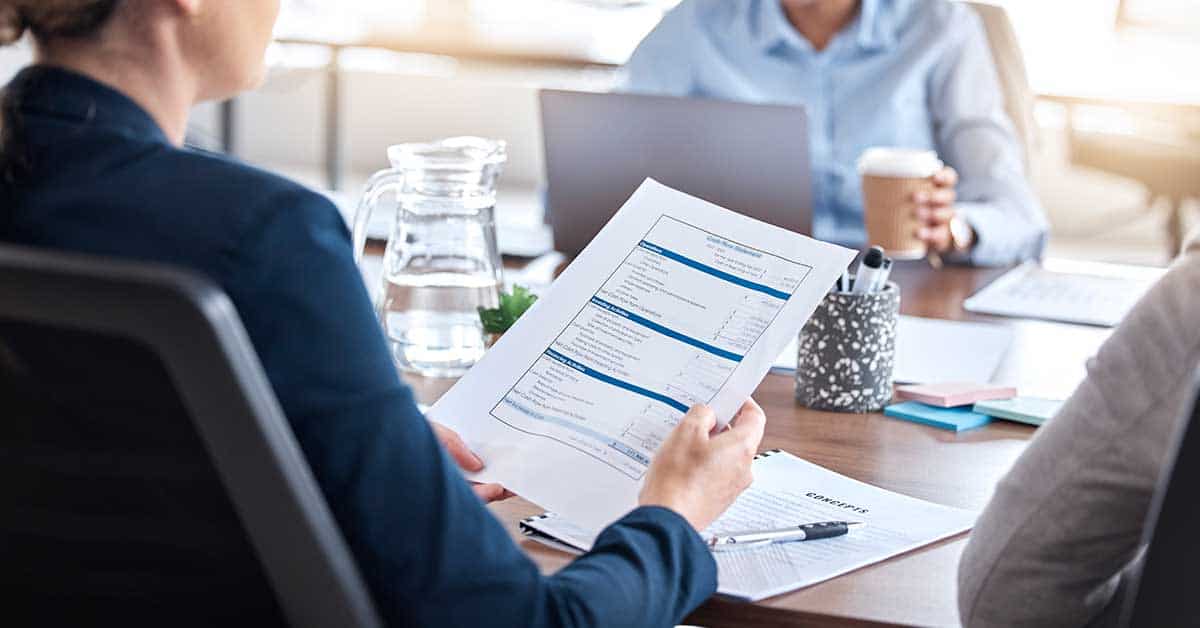 In a meeting room, a person reviewing compliance requirements is seated at a table. Nearby, one individual focuses on their laptop while another sips from a coffee cup. A glass water pitcher stands in the background, adding a touch of elegance to the setting.