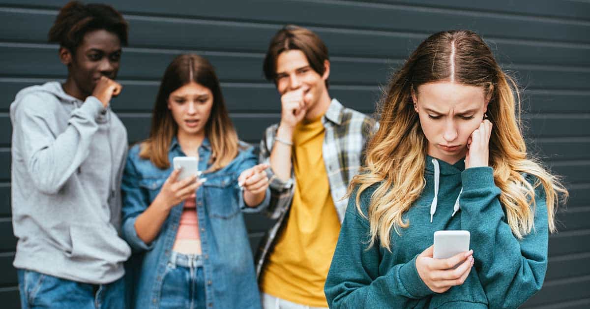 A girl looks upset while checking her phone, possibly after seeing a deepfake, as three other teenagers stand behind her, reacting with surprised and amused expressions.
