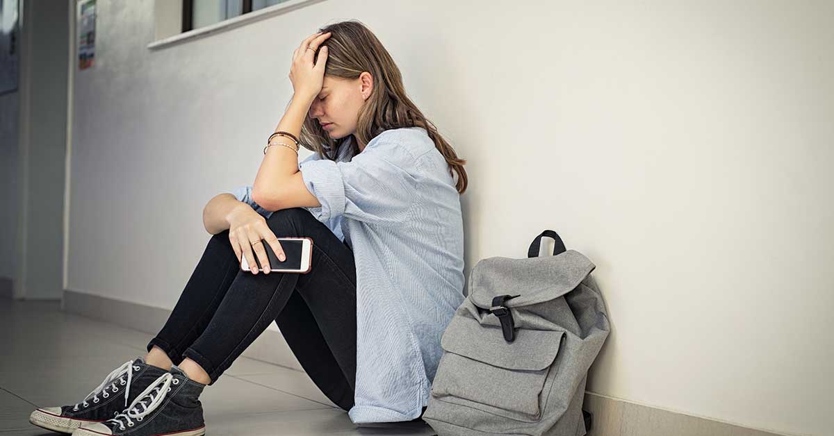 A young woman sits on the floor against a wall, holding her head in one hand and a phone in the other, with a backpack beside her. She appears distressed, possibly facing exploitation.