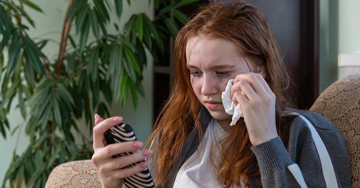 A young woman sits on a couch, wiping tears from her face with a tissue while looking at her phone, realizing that ignorance is not bliss in the age of digital exploitation.