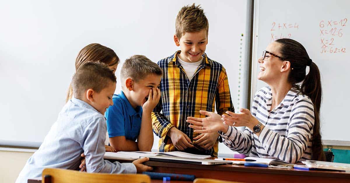 A teacher and four children are gathered around a desk in a classroom, smiling and talking, with books and markers on the table, enjoying an engaging education experience together.