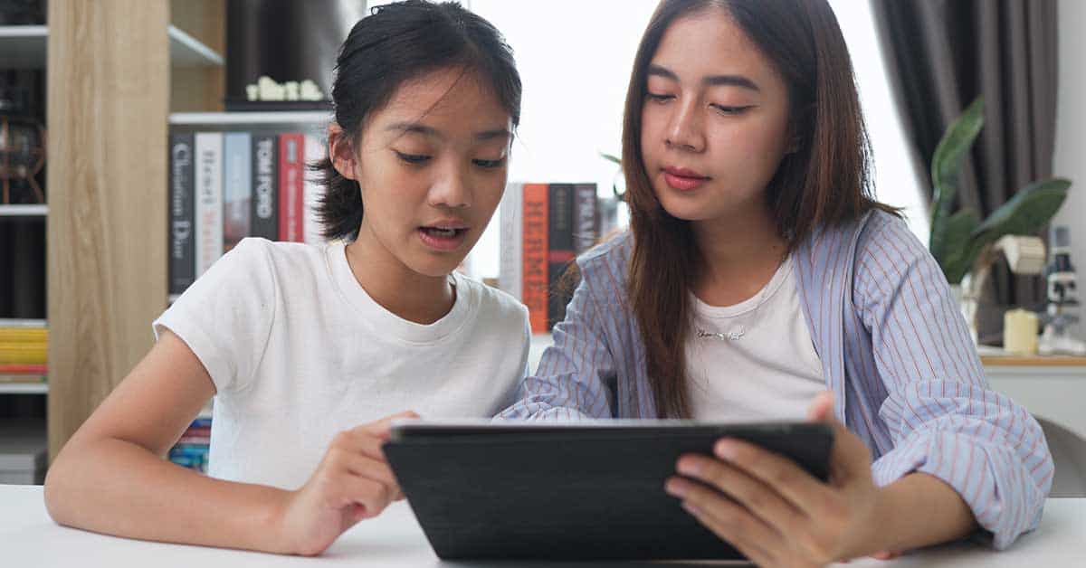 Two students sit at a table, looking at a tablet together. Books are visible on a shelf in the background.