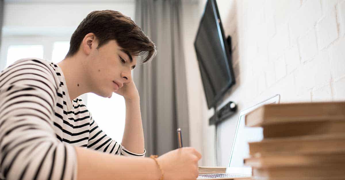Young academic in a striped shirt sits at a desk, writing in a notebook, with a laptop and stacked books nearby.