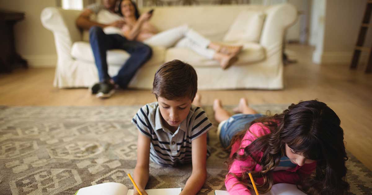 Two children lie on a rug drawing with pencils, while two adults sit on a white couch in the background—an inviting scene perfect for encouraging enrollment in family programs.