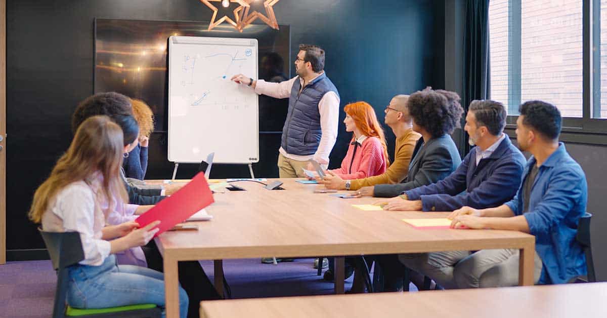 A man stands at a whiteboard presenting a diagram to a group of people seated around a conference table in a meeting room, creating an atmosphere similar to an engaging school classroom discussion.