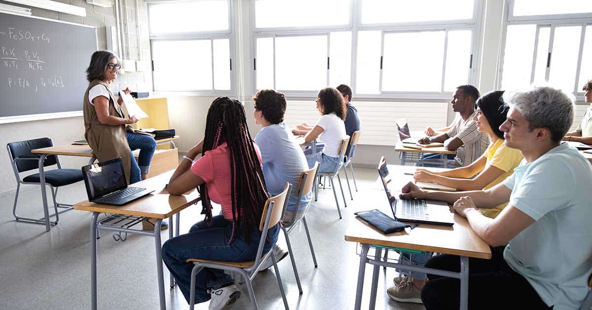 A teacher stands at the front of a classroom giving a literacy lesson to students seated at desks, some taking notes and using laptops.