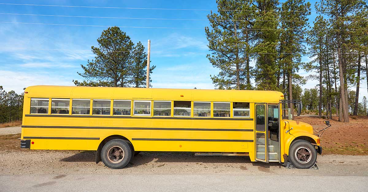 A yellow BIE school bus with a flat front tire is parked on the side of a rural road near trees under a blue sky.