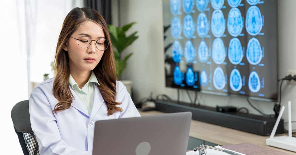 A woman in a lab coat works on a laptop at a desk, analyzing brain scan images with the help of AI, displayed on a large screen in the background.