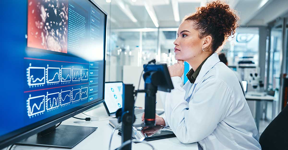 A scientist in a lab coat reviews data charts and AI-analyzed microscopic images on a large computer monitor at a laboratory workstation.