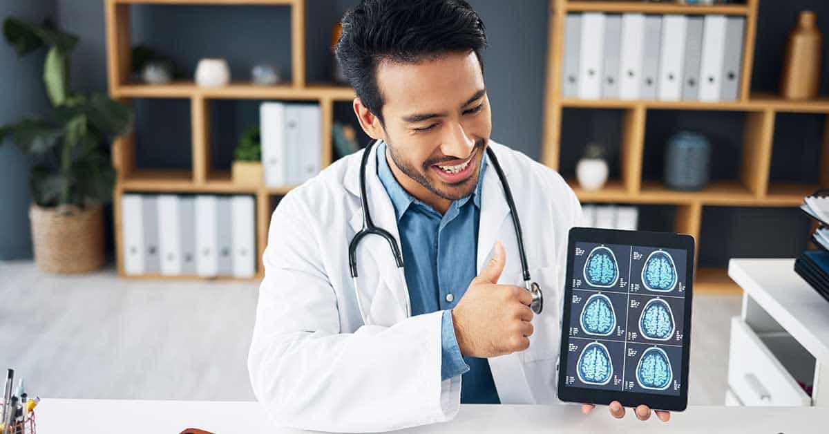 A doctor in a white coat and stethoscope smiles and gives a thumbs-up while showing AI-powered brain scan images on a tablet.