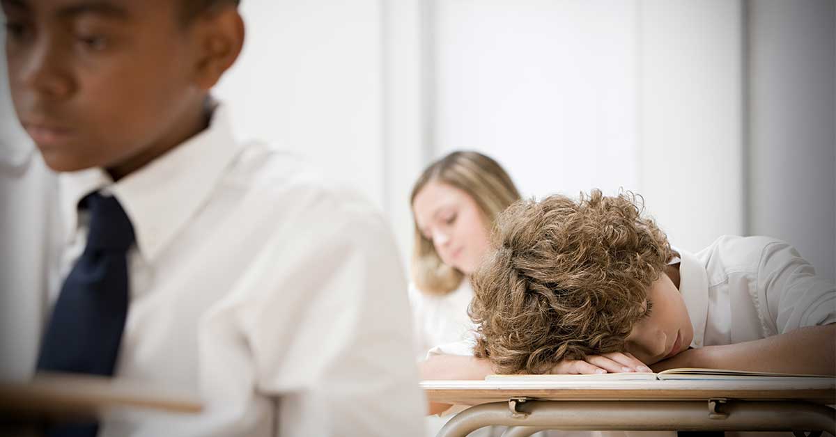 A student with curly hair sleeps on a desk in a classroom, facing the consequences of exhaustion, while other students sit nearby—one focused and one looking forward.