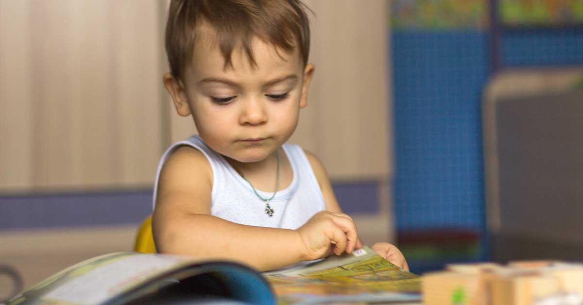 A toddler in a white tank top is focused on flipping through an open book while seated indoors, nurturing early literacy skills and setting the foundation for kindergarten readiness.