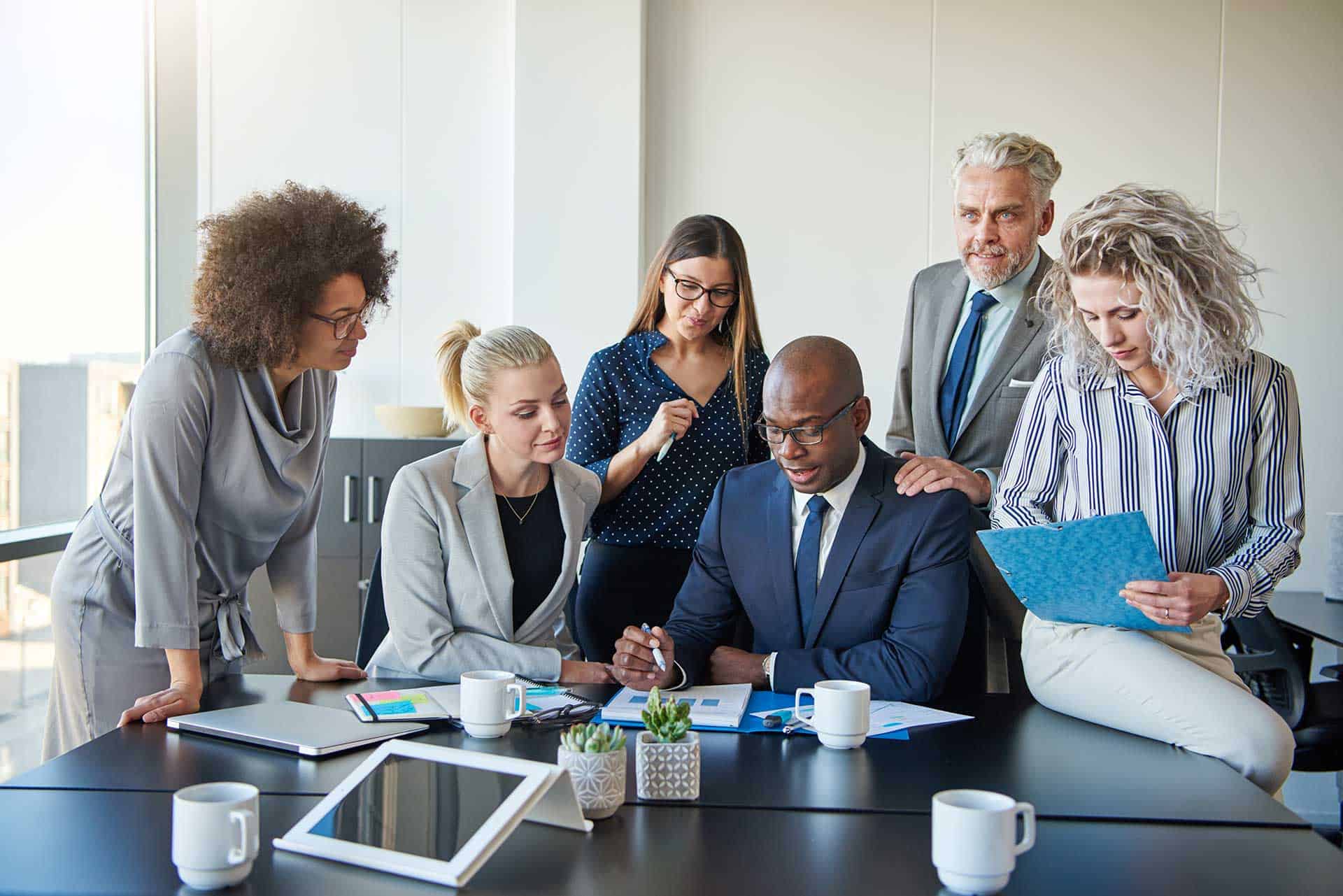 A diverse group of business professionals, committed to excellence, gathers around a conference table, discussing documents and using digital devices.