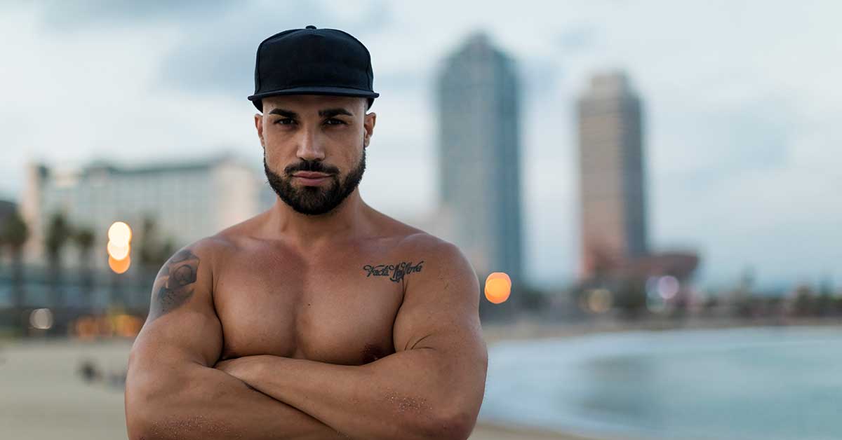 A muscular, shirtless man wearing a black cap stands with arms crossed on a beach, exuding confidence—a stark contrast to the incel stereotypes—while blurred city buildings loom in the background.