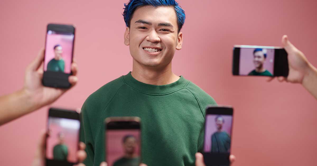 A person with blue hair and a green sweater smiles confidently while several hands hold up smartphones, capturing their moment of activism against a vibrant pink background.