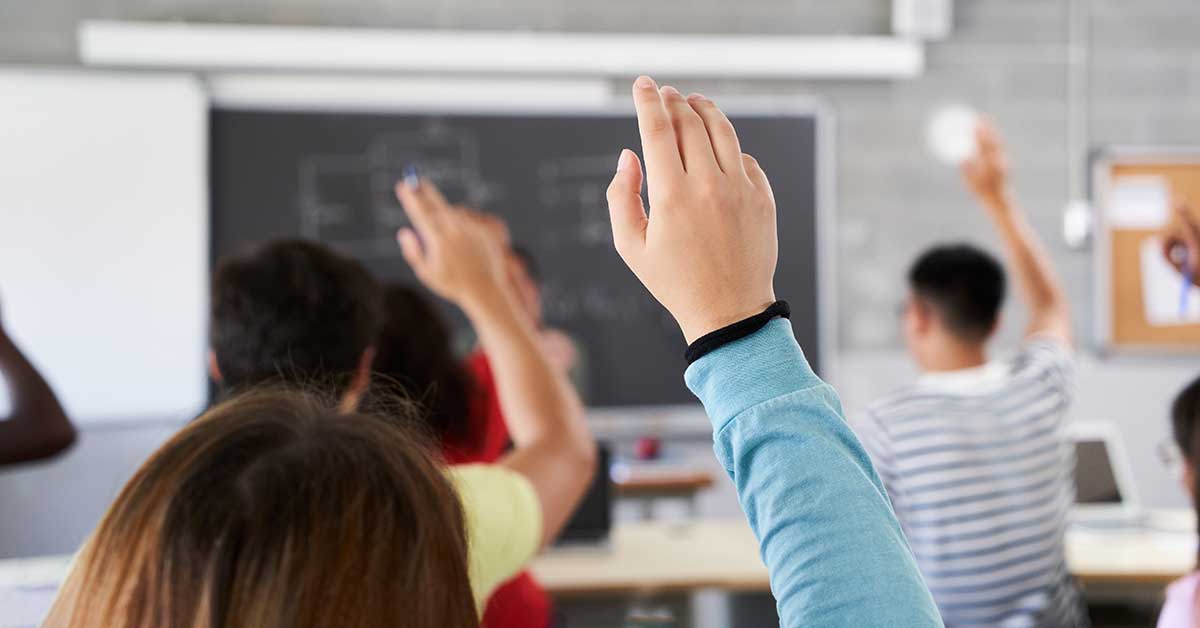 Several students sitting in a classroom raise their hands, facing a chalkboard at the front of the room, as they engage in a lively discussion about the economy and its impact on schools.