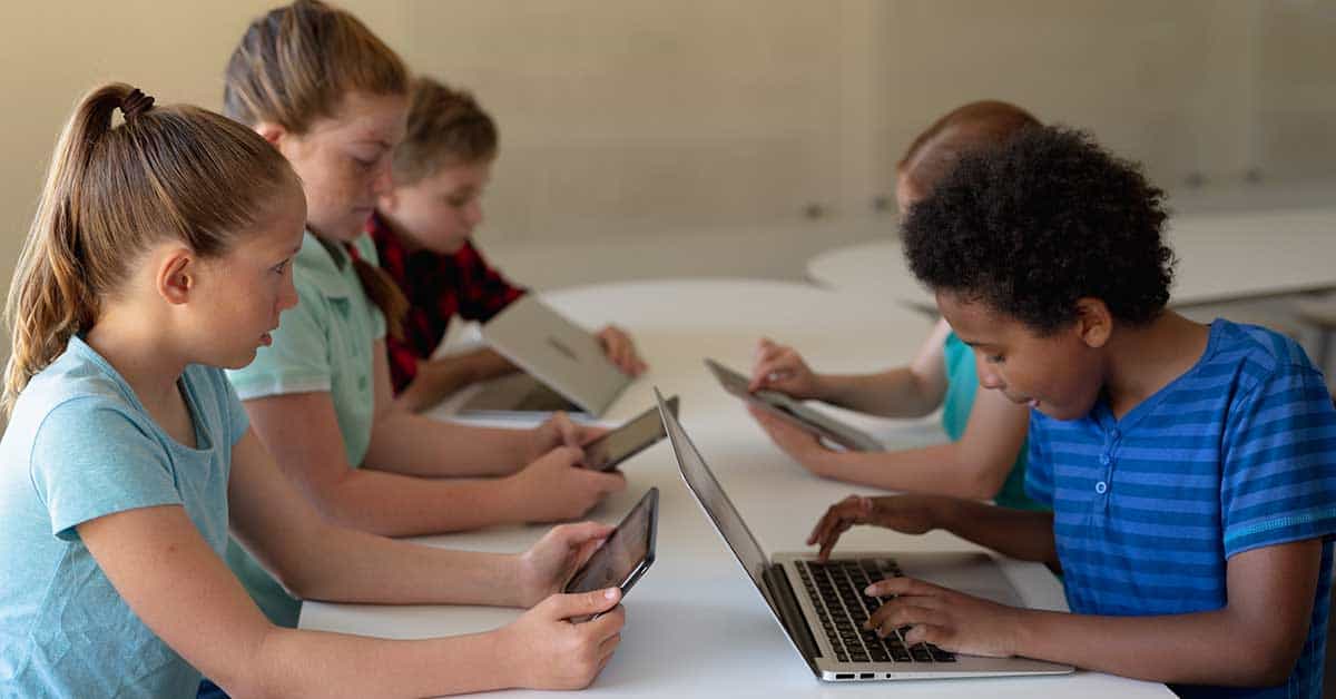 Children adapt to the technological revolution, focused on using tablets and laptops while sitting at a table in a classroom setting.